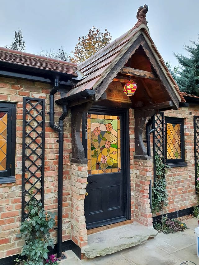 Bespoke stained-glass front door set in brickwork beneath a clay-tiled timber porch on a garden room in Surrey