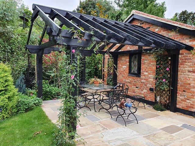 Black timber pergola shading a sandstone patio dining area with climbing roses beside a brick garden room in Surrey