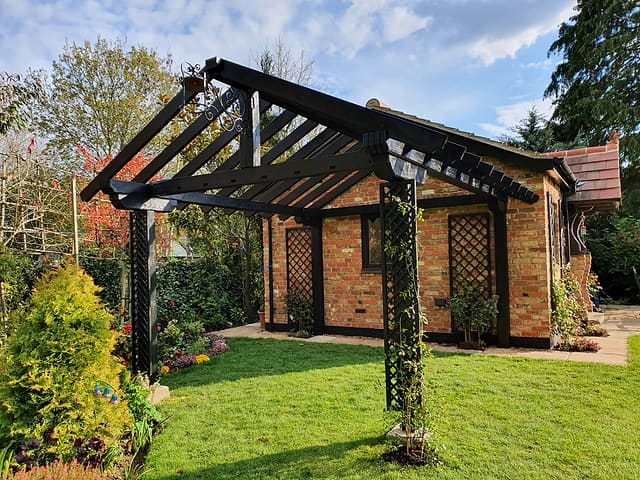 Black timber pergola with trellis posts beside a brick garden room under a blue sky in a Surrey garden